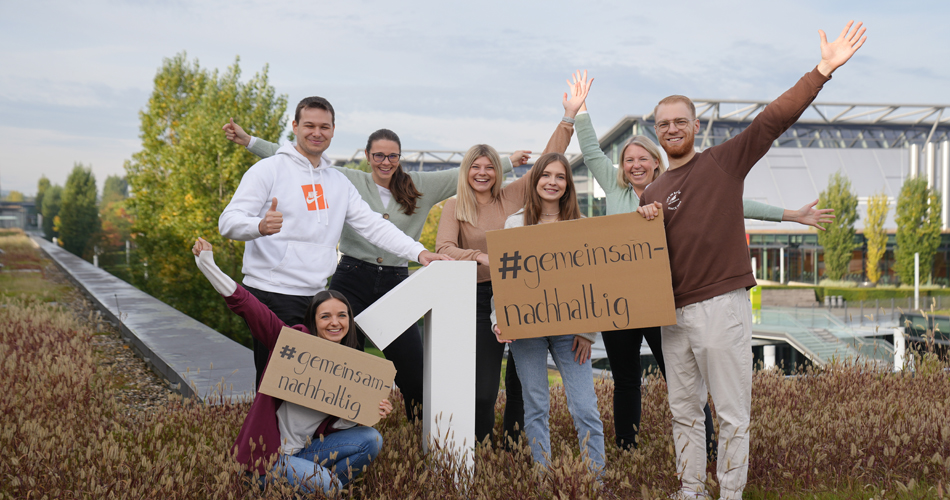 Eine Gruppe von sechs Personen steht lächelnd im Freien und hält Schilder mit der Aufschrift #gemeinsamnachhaltig in die Kamera, während sie freudig die Arme hochheben. Im Hintergrund sind Bäume und das Messegelände zu sehen.